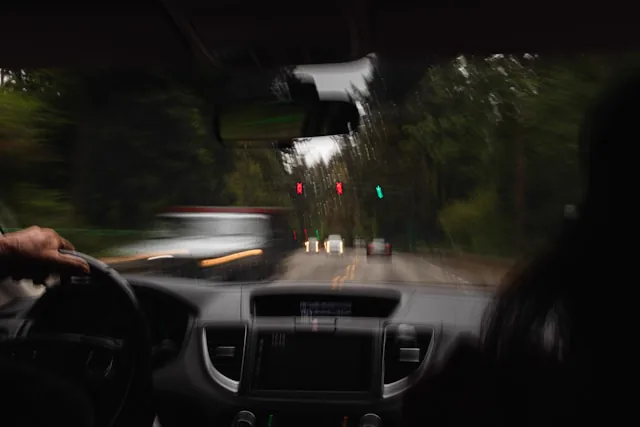 Verschwommene Sicht durch die Windschutzscheibe bei Dämmerung, Hand am Lenkrad, Ampel und Gegenverkehr voraus