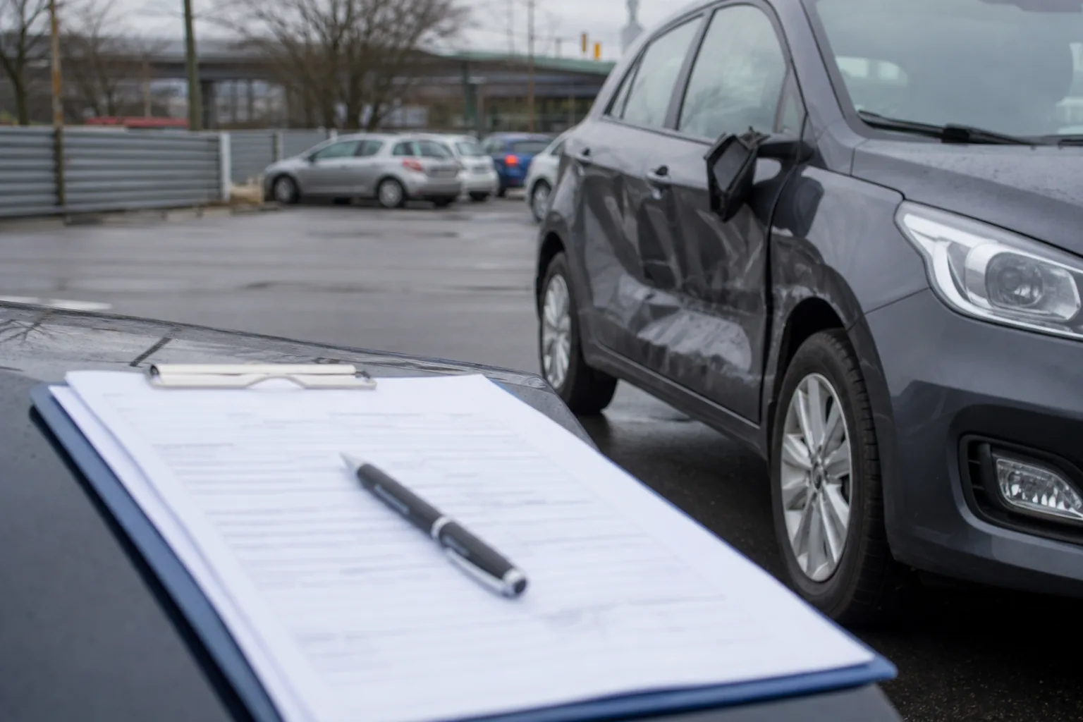 Klemmbrett mit Schadenformular auf einem Autodach, dahinter ein grauer Wagen mit eingedrückter Fahrzeugseite