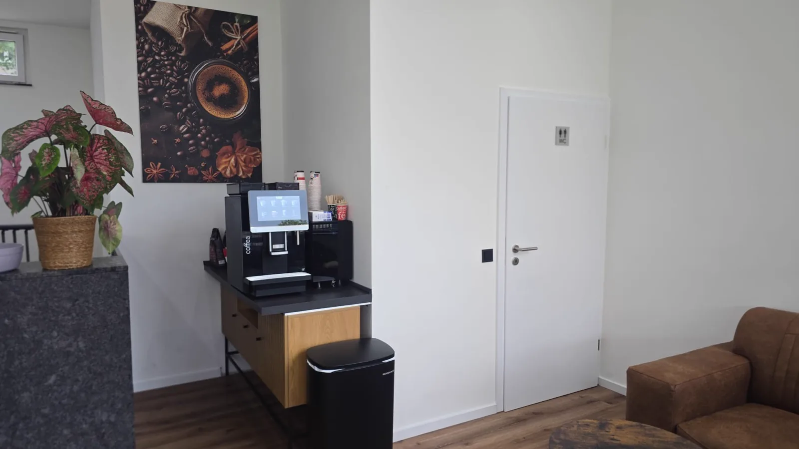 Wartebereich mit Kaffeevollautomat auf dem Sideboard, Sessel und großem Kaffeebild an der Wand